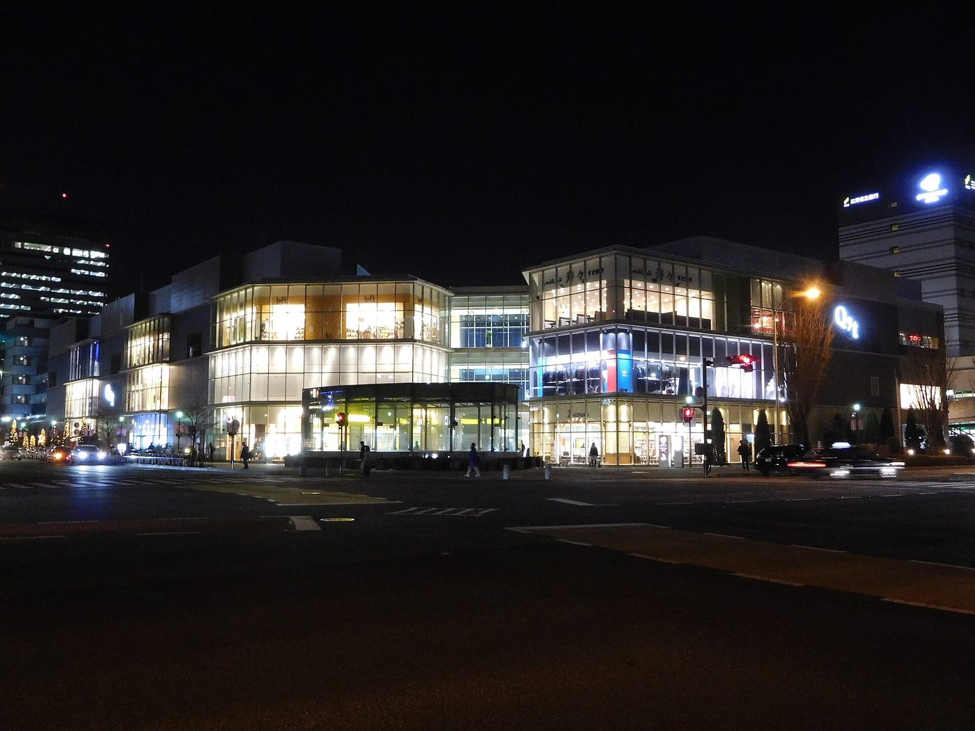 夜のつくば駅前。内側から灯りの漏れるQ’tとクレオの建物、濡れた路面と信号